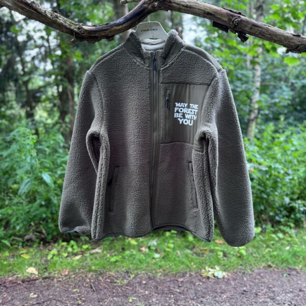 Fleecetrøje med lynlås i gråbrun fleece med “May the Forest Be With You”-tryk på brystet - vist hængende på en gren langs en skovsti