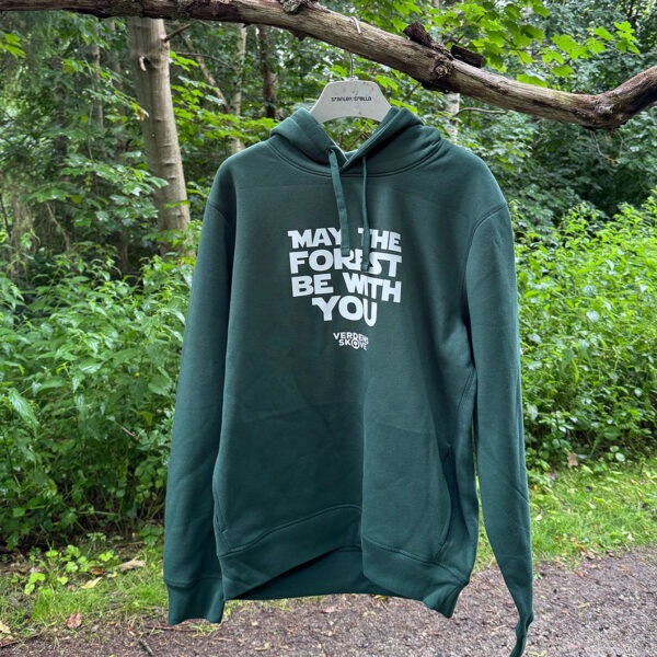 Grøn hoodie med hvidt “May the Forest Be With You”-tryk på forsiden - set hængende på en gren langs en sti i skoven
